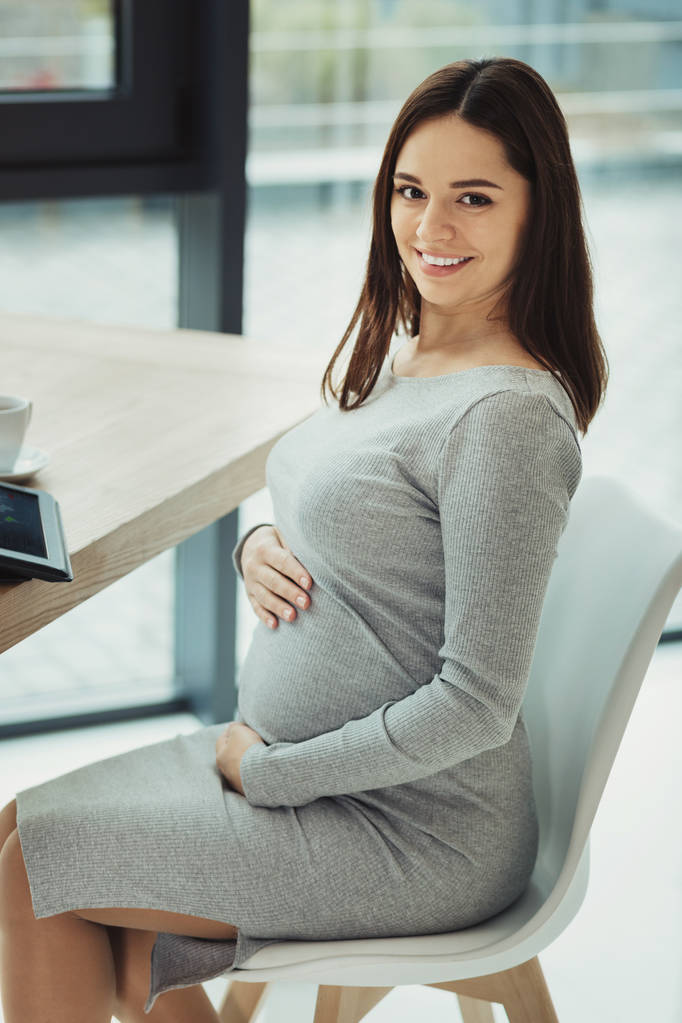 Miellyttävä odotus. Kaunis nuori raskaana oleva sihteeri pehmeästi koskettaa hänen vatsansa istuessaan pöydässä nykyaikaisessa toimistossaan
 - Valokuva, kuva
