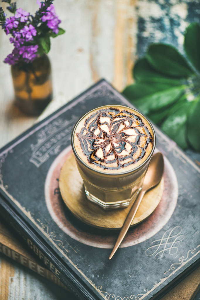 Café con leche con patrón de salsa de chocolate en vaso alto sobre mesa rústica con fondo de libros
 - Foto, imagen