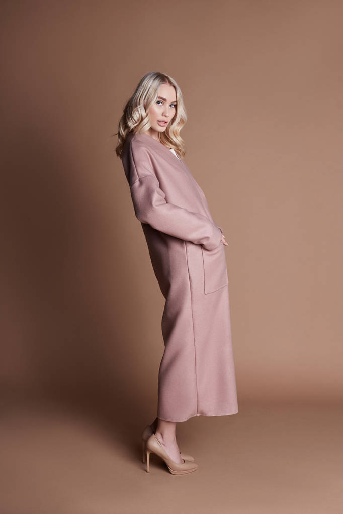 Mooie blonde vrouw poseren in een roze jas op een beige achtergrond. Modeshow van kleding, vrouw met perfecte figuur, lange haren. Trendy modieuze herfst jas op lichaam van het meisje - Foto, afbeelding