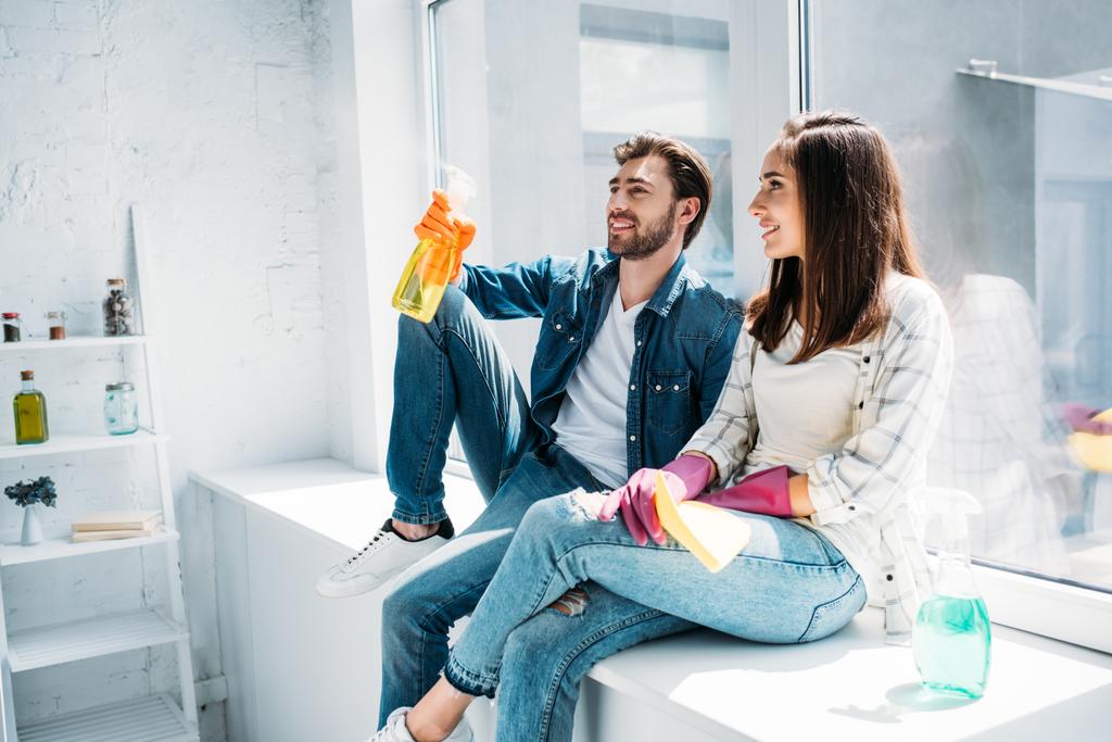 Paar ruht sich nach Putzen auf Fensterbank aus und hat Spaß mit Sprühflasche in Küche - Foto, Bild