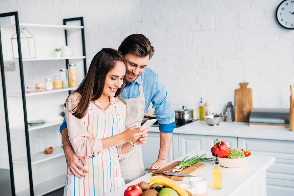 пара обнимает и смотрит на смартфон во время приготовления пищи на кухне
 - Фото, изображение