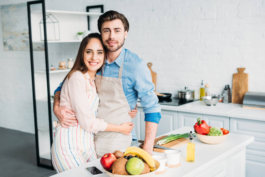 ζευγάρι σε ποδιές αγκάλιασμα και βλέπουν τα φωτογραφικών μηχανών στην κουζίνα - Φωτογραφία, εικόνα
