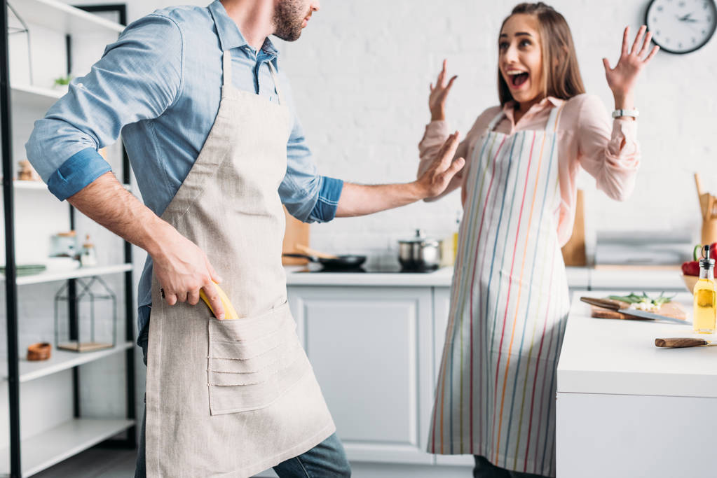 excited girlfriend standing with hands up and boyfriend holding banana gun in kitchen - Photo, Image