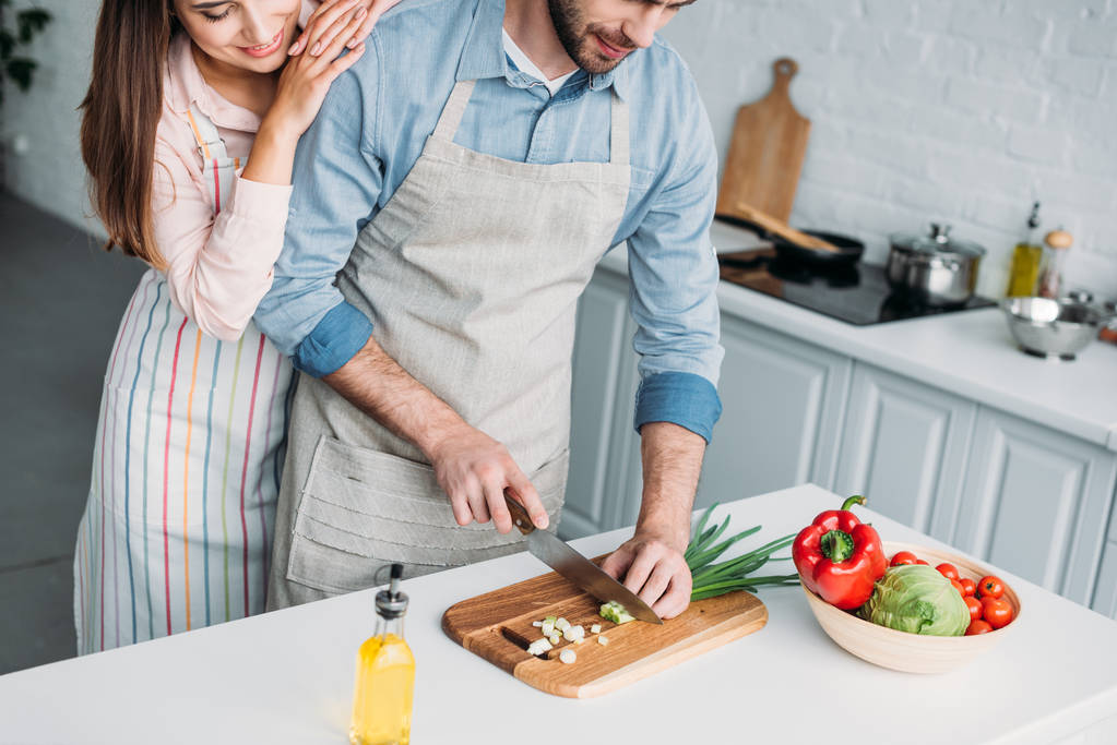 rajattu kuva poikaystävä leikkaa vihanneksia ja tyttöystävä nojaa häneen keittiössä
 - Valokuva, kuva