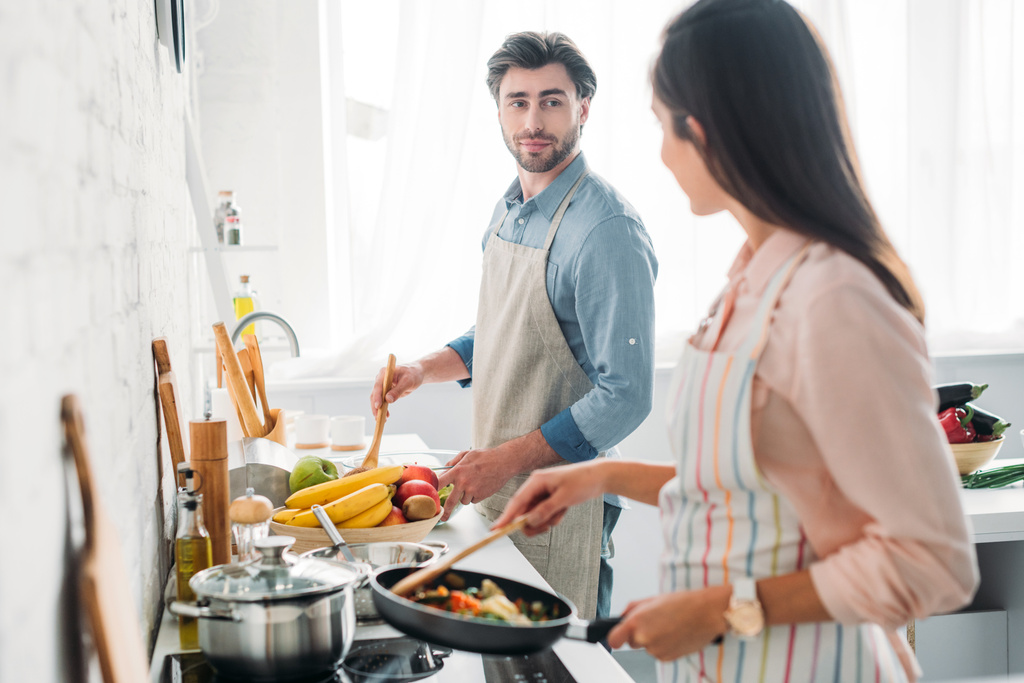 girlfriend frying vegetables on frying pan in kitchen and looking at boyfriend - Photo, Image