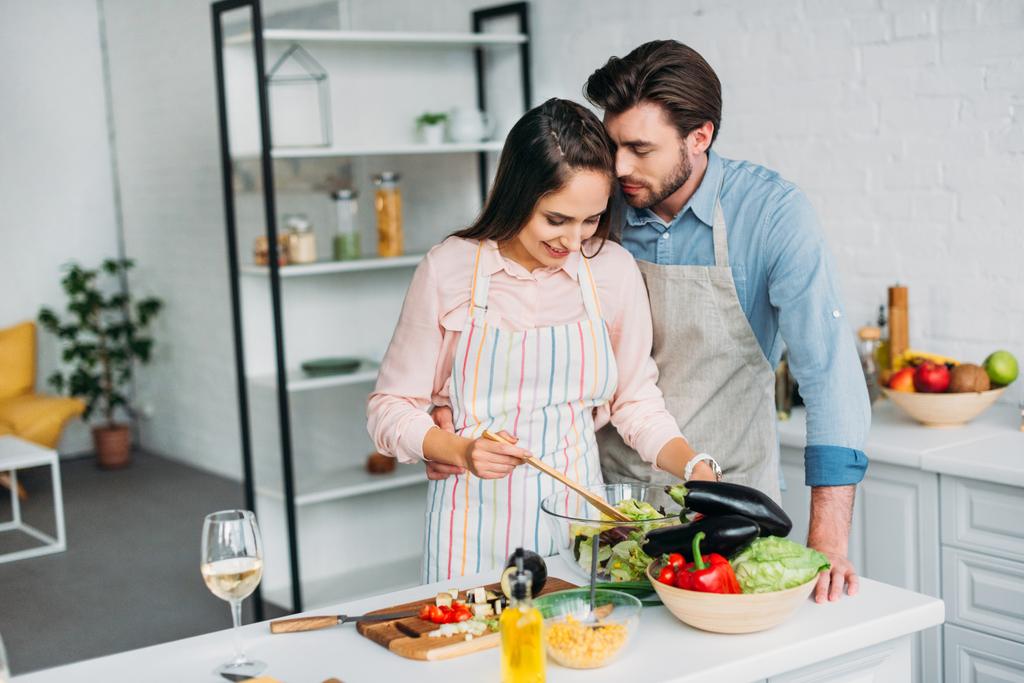 Freundin kocht und mixt Salat in Küche und Freund umarmt sie - Foto, Bild