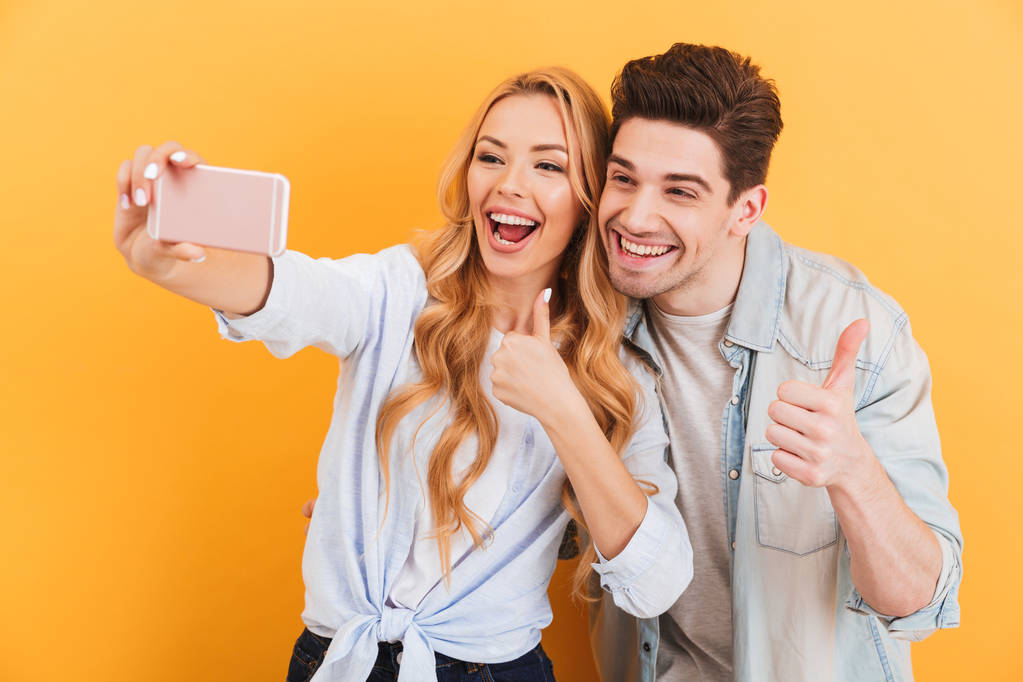 Портрет счастливой пары мужчины и женщины, делающей селфи на мобильном телефоне, в то время как жесты на камеру большие пальцы вверх изолированы на желтом фоне
 - Фото, изображение
