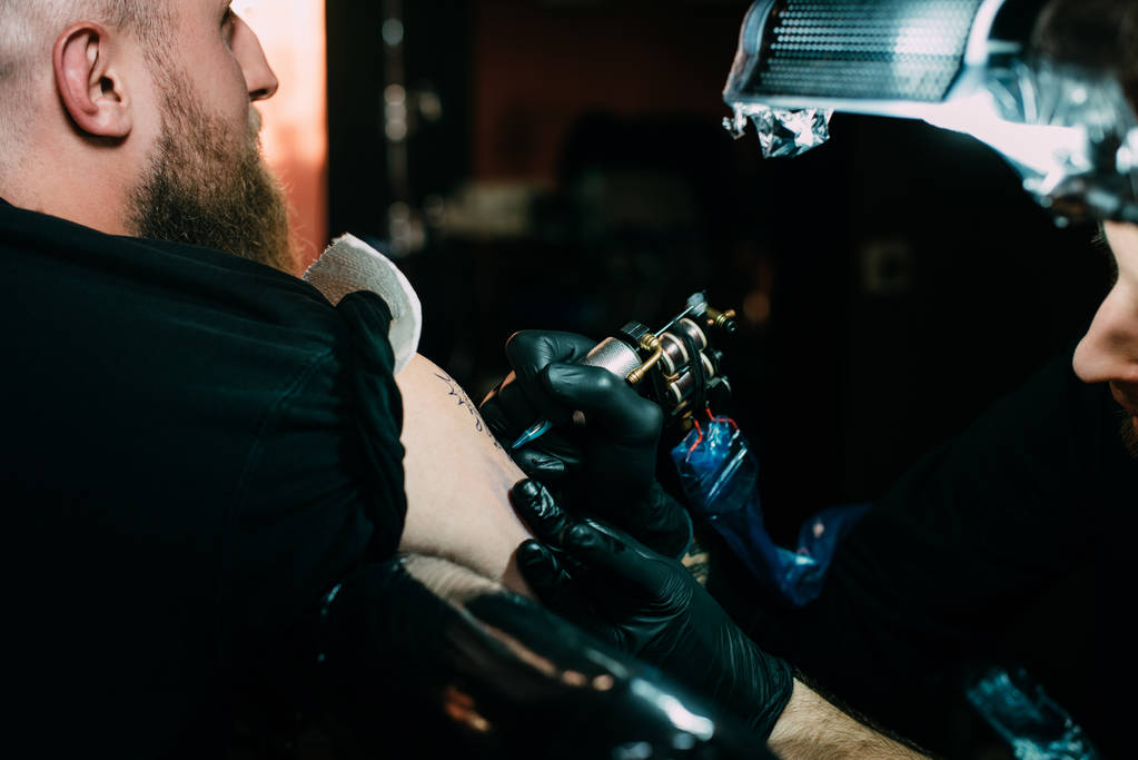 селективный фокус татуировщика в перчатках с тату-машиной, работающей на татуировке на плече в салоне
 - Фото, изображение