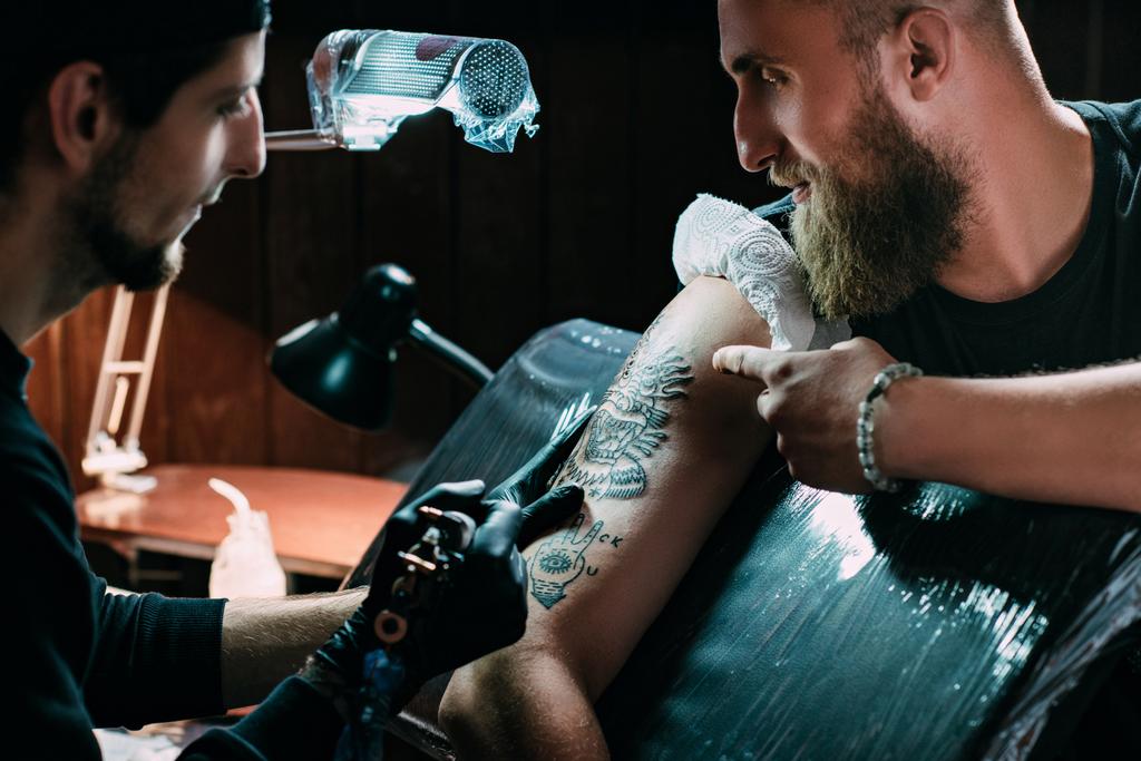 Μερική άποψη του καλλιτέχνη τατουάζ στα γάντια με τατουάζ μηχανή που λειτουργεί στα τατουάζ στον ώμο στο σαλόνι - Φωτογραφία, εικόνα