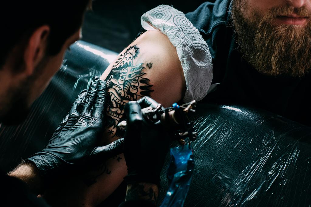 bijgesneden schot van tatoeëerder in handschoenen werken op tatoeage op schouder in salon - Foto, afbeelding