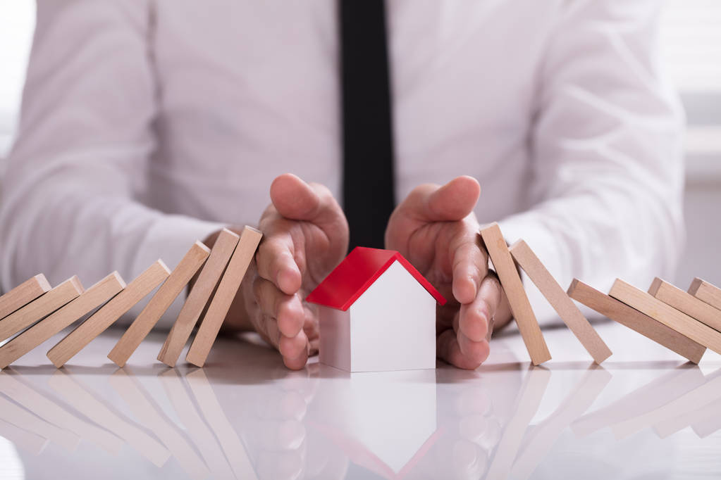 Close-up Of A Businessperson's Hand Protecting House Model From Falling Wooden Blocks On White Desk - Photo, Image