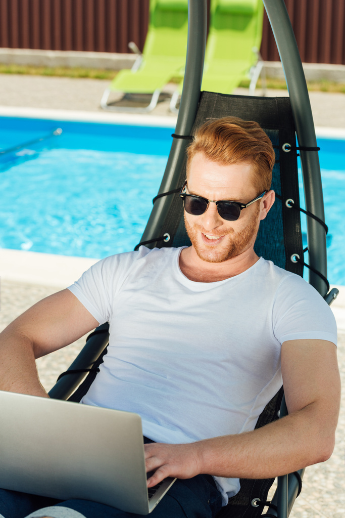 jovem bonito sentado na espreguiçadeira em frente à piscina e trabalhando com laptop
 - Foto, Imagem