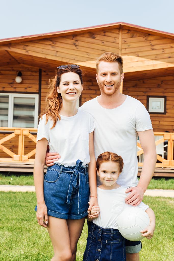 happy young family with volleyball ball standing in front of wooden cottage - Photo, Image