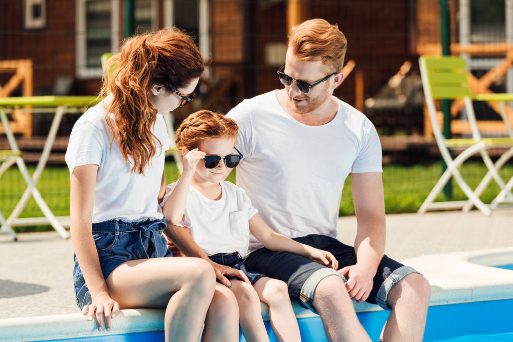 Junge Familie in weißen T-Shirts und Sonnenbrille sitzt gemeinsam am Pool - Foto, Bild