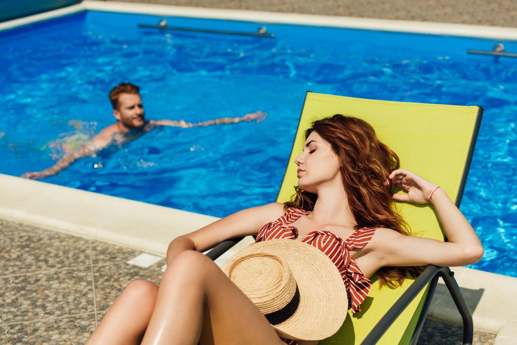 jonge vrouw ontspannen op een ligstoel terwijl haar vriendje zwemmen in zwembad op achtergrond - Foto, afbeelding