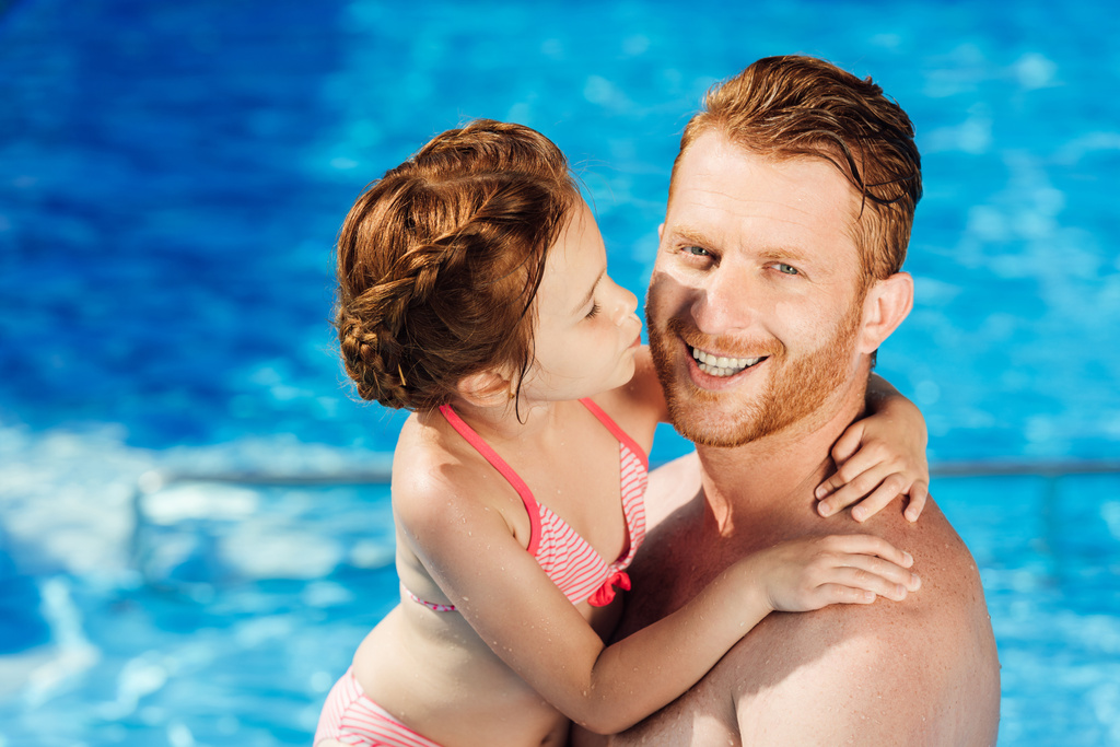 glücklicher Vater und Tochter umarmen sich im Schwimmbad und blicken in die Kamera - Foto, Bild