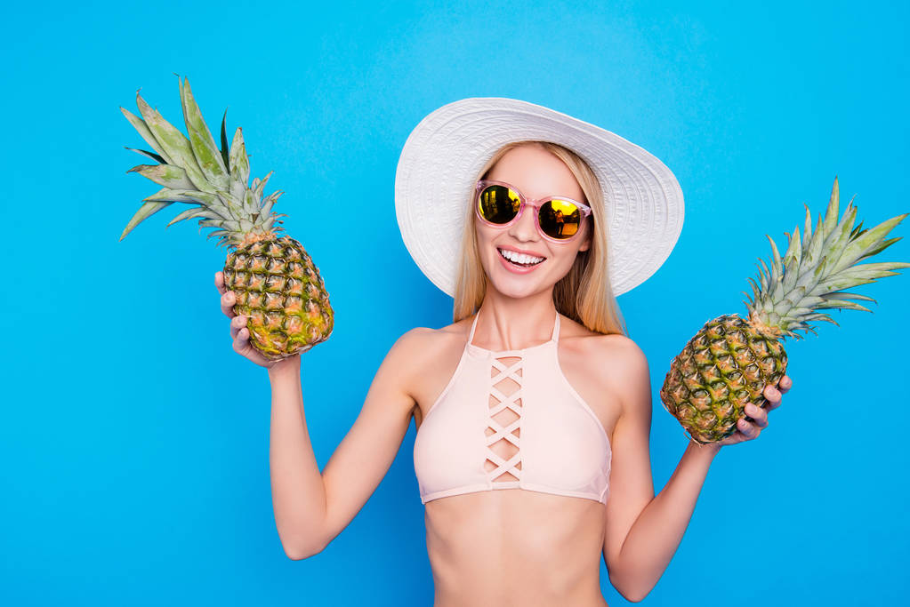 Rózsaszín melltartó nyári szem szemüveg két friss ananász tartja kezében, nézi a kamera elszigetelt világos kék háttér-vidám szép lány portréja - Fotó, kép