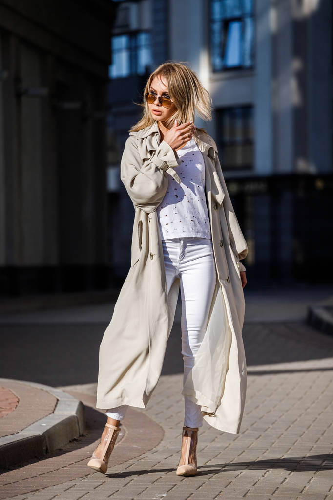 Внешний портрет блондинки в бежевом пиджаке, белой футболке и белых брюках, стоящей на улице с размытым зданием на заднем плане
 - Фото, изображение