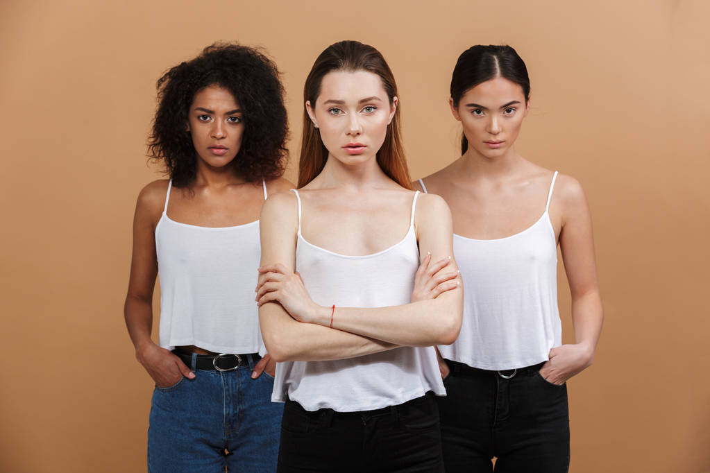 Obraz ze tří mezinárodních žen: Kavkazský, africké americké a asijské dívky v neformálním oblečení s přísným pohledem, samostatný béžové pozadí při pohledu na fotoaparát - Fotografie, Obrázek
