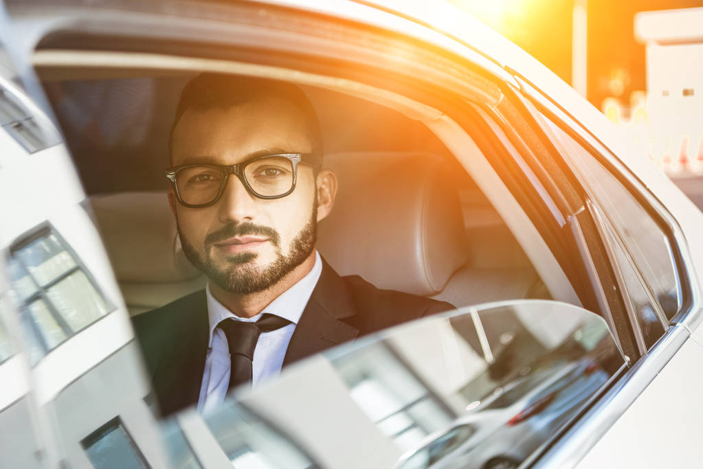 handsome businessman sitting in car and looking at camera during sunset - Photo, Image