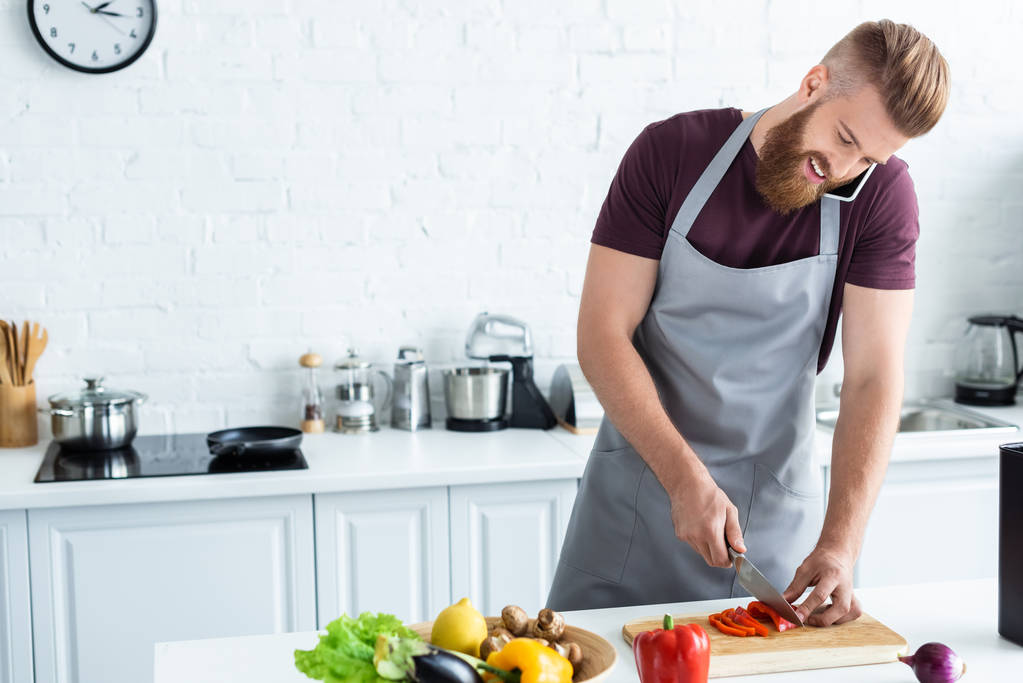 gülümseyen genç adam sebze kesme ve yanında smartphone mutfakta konuşmak önlük sakallı - Fotoğraf, Görsel