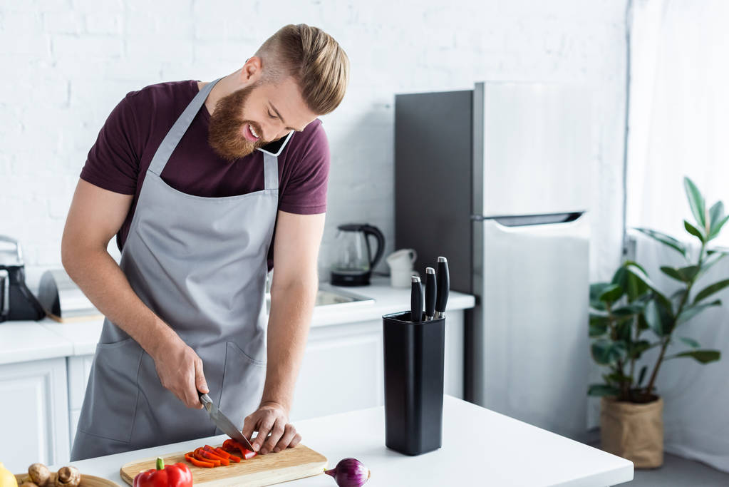 handsome smiling bearded man in apron talking by smartphone and cutting vegetables in kitchen   - Photo, Image