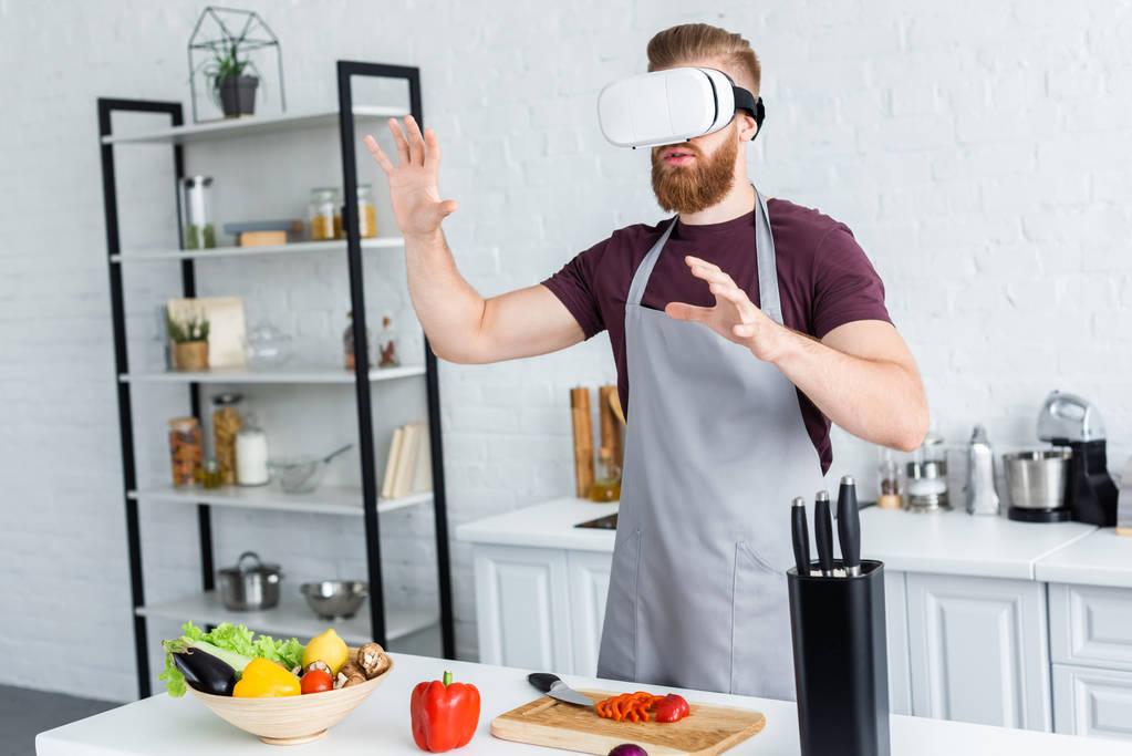 Junger Mann im Kittelschürzen mit Virtual-Reality-Headset beim Kochen in der Küche    - Foto, Bild