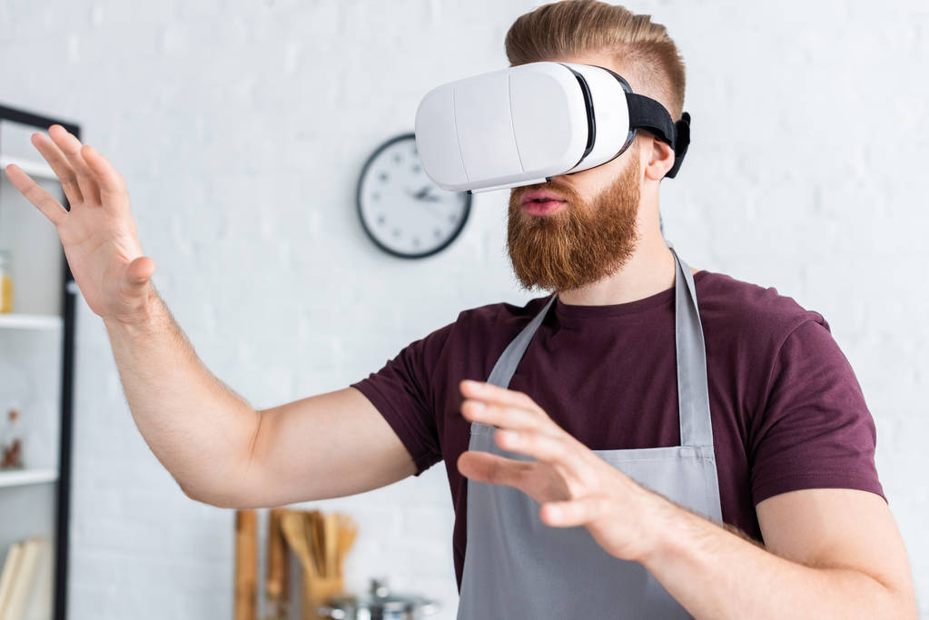 bearded young man in apron using virtual reality headset in kitchen  - Photo, Image