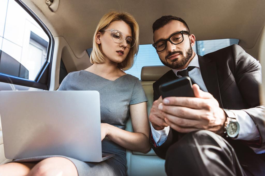 low angle view of handsome businessman showing something on smartphone to assistant in car - Photo, Image