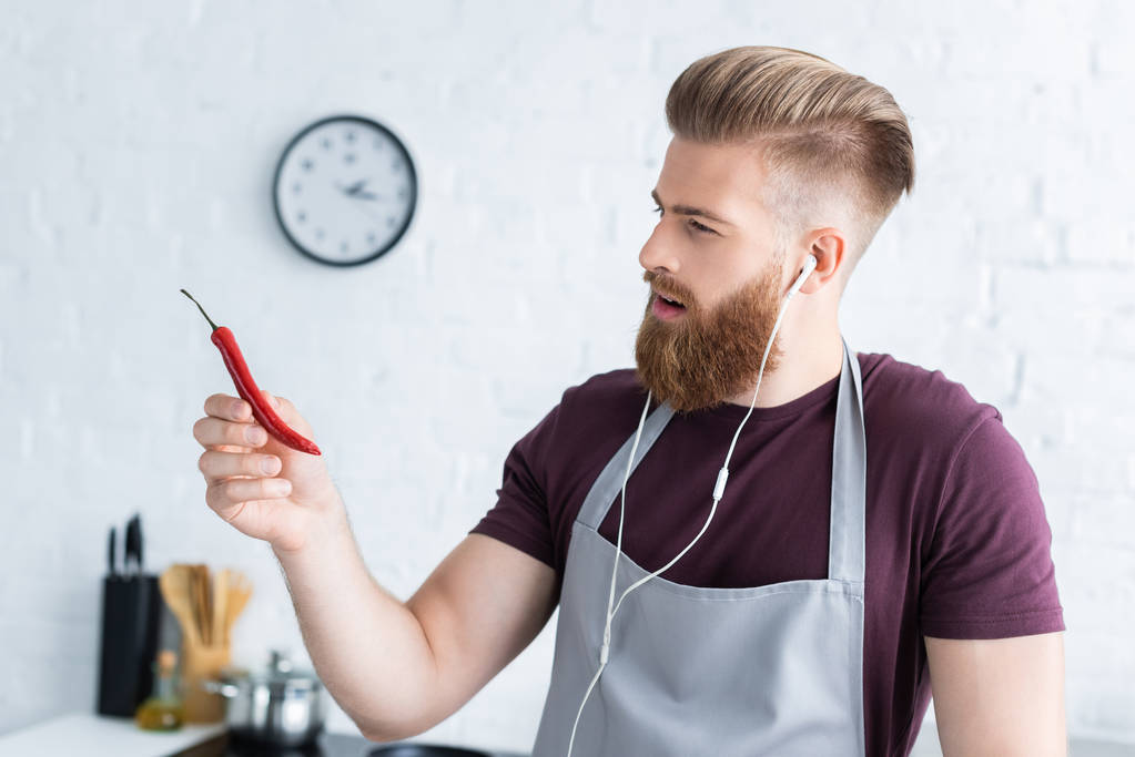 handsome bearded young man in apron and earphones holding chili pepper in kitchen - Photo, Image