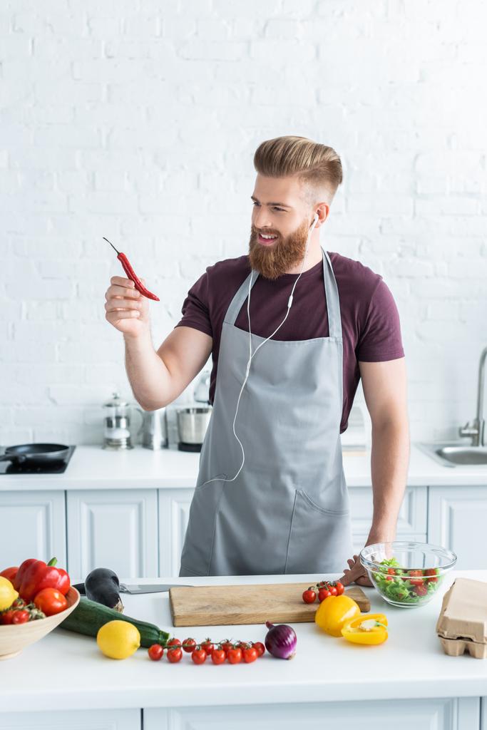 yakışıklı gülümseyen adam kulaklık müzik dinleme ve mutfakta yemek yaparken chili biber tutan önlük sakallı - Fotoğraf, Görsel