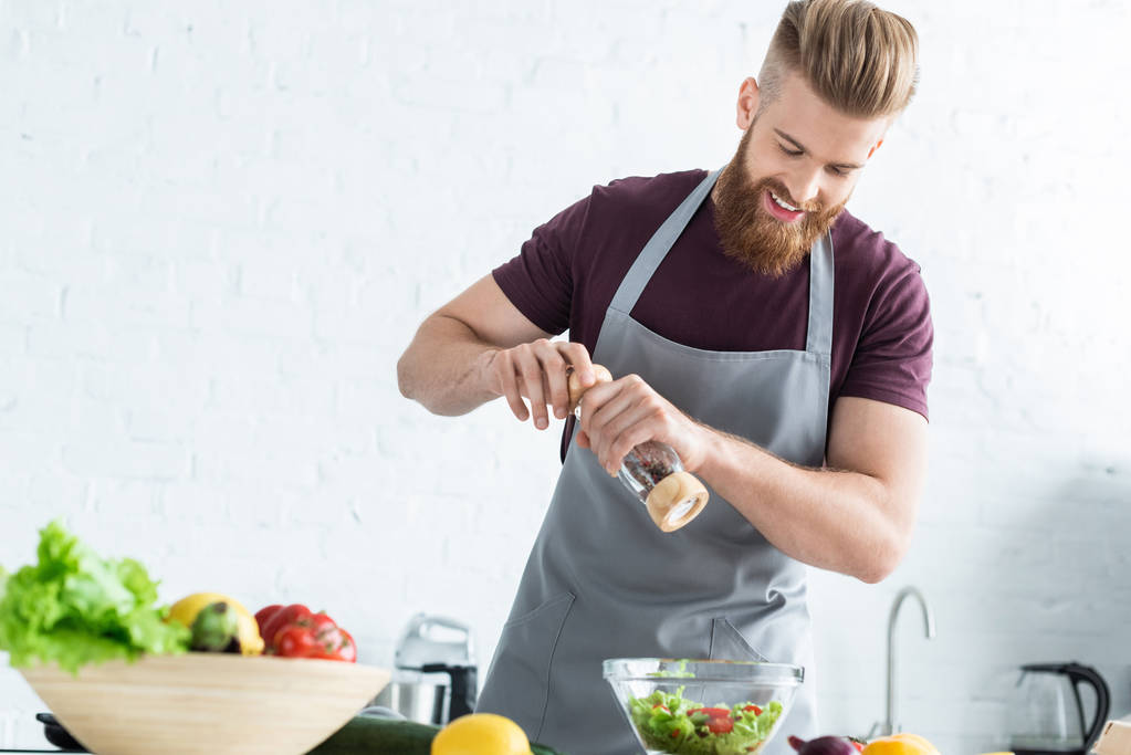 handsome smiling young man in apron spicing vegetable salad - Photo, Image