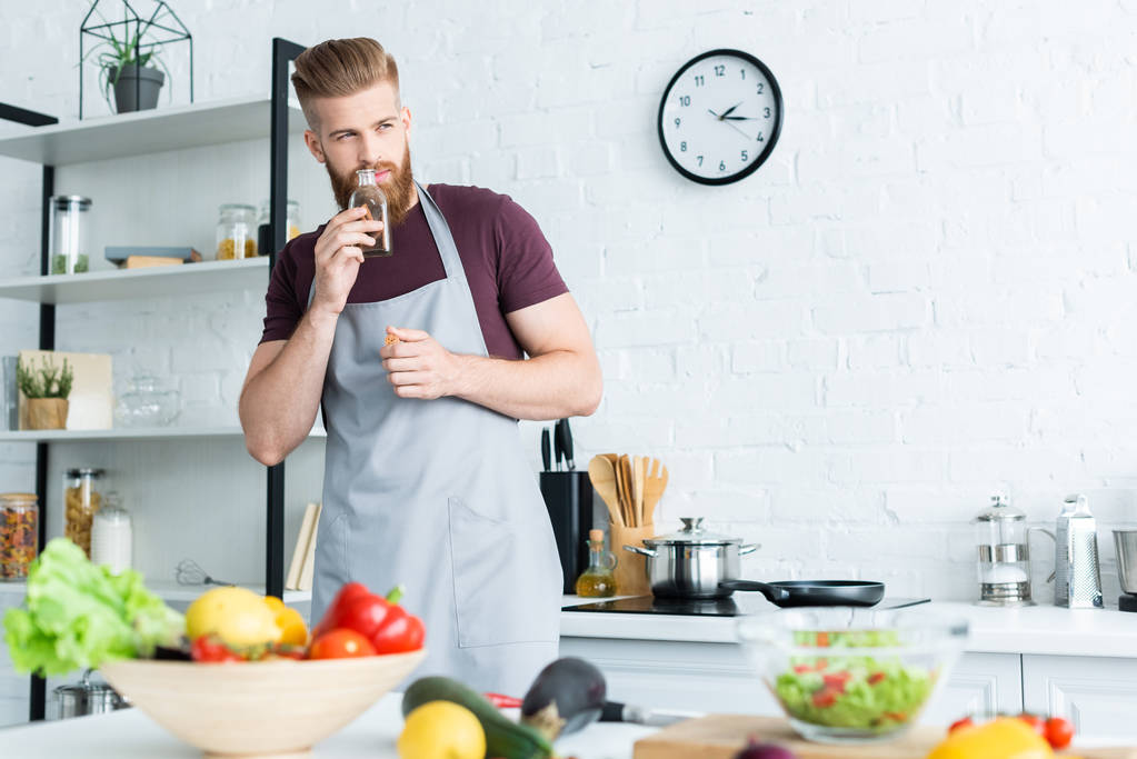 giovane bell'uomo barbuto in grembiule guardando lontano mentre cucinava in cucina
 - Foto, immagini
