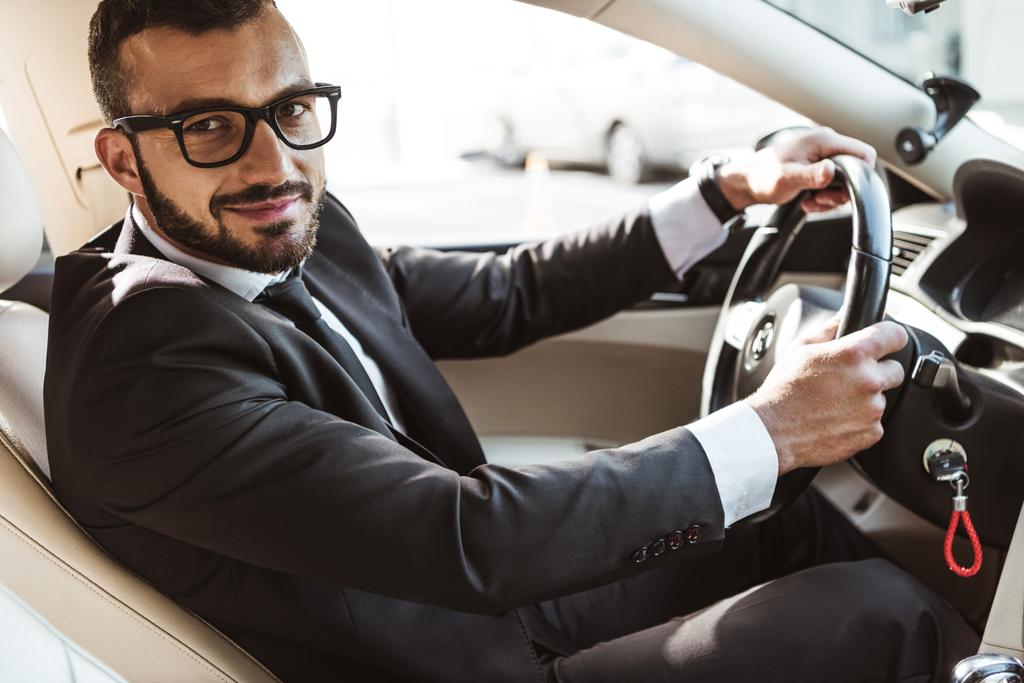 Вид сбоку красивого водителя в костюме, управляющего машиной и смотрящего в камеру
 - Фото, изображение