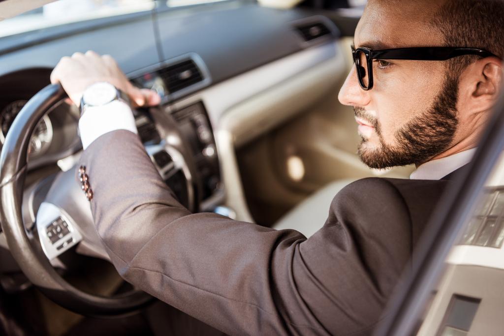 Вид сбоку красивого водителя в костюме и очках за рулем автомобиля
 - Фото, изображение