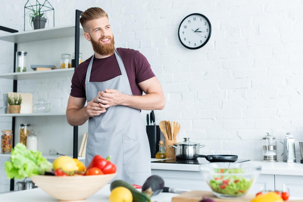 przystojny uśmiechający się Brodaty mężczyzna w fartuch, którzy szukają drogi podczas gotowania w kuchni - Zdjęcie, obraz