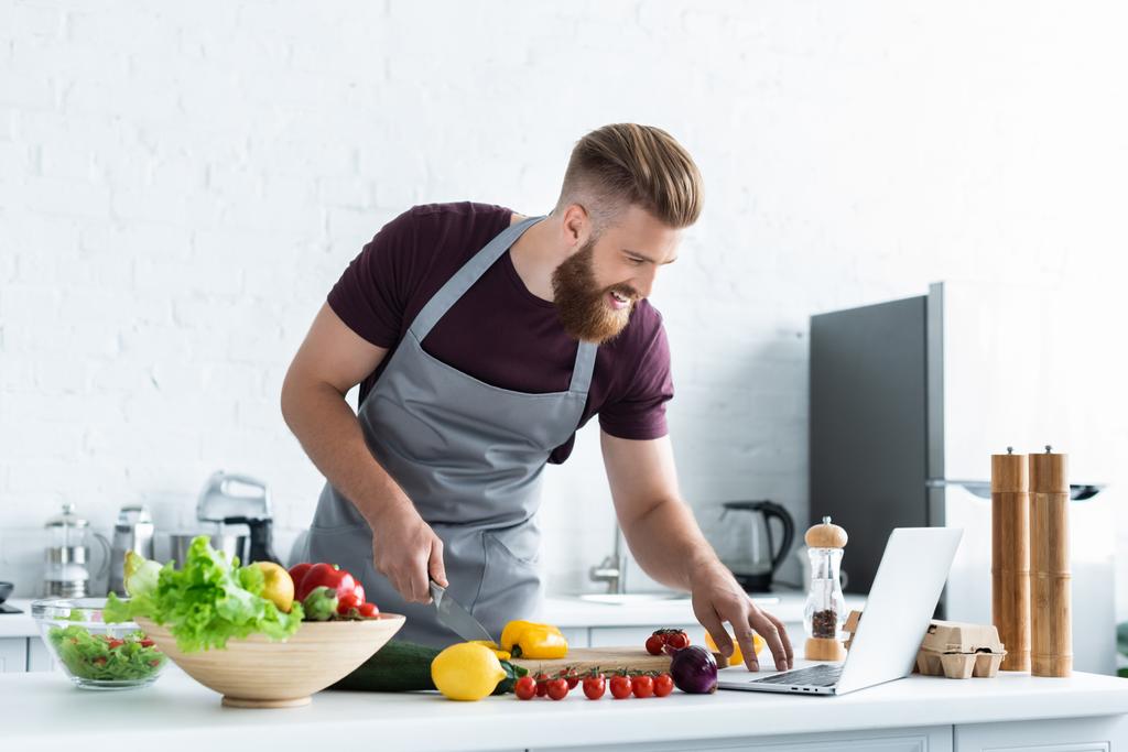 mosolygó szakállas férfi-kötény használ laptop és főzés zöldségsaláta  - Fotó, kép