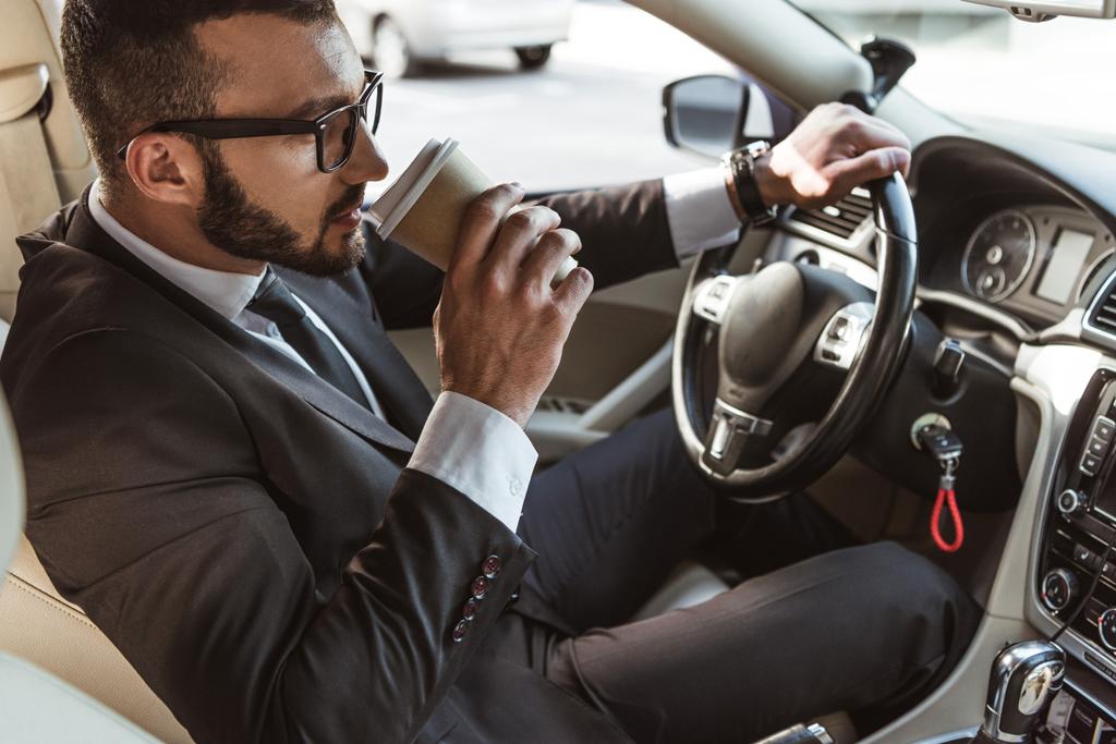 knappe chauffeur in pak rijdende auto en drinken koffie in papier beker - Foto, afbeelding