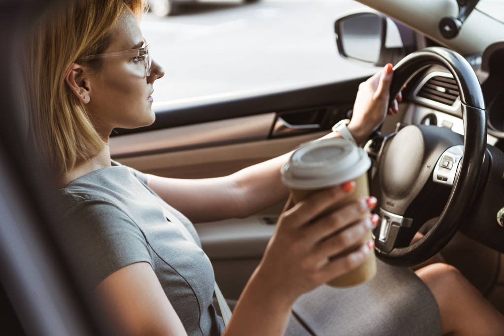 Zijaanzicht van aantrekkelijke zakenvrouw auto rijden en koffie houden in papier beker - Foto, afbeelding