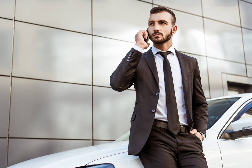 knappe zakenman praten door smartphone en permanent in de buurt van de auto op straat - Foto, afbeelding