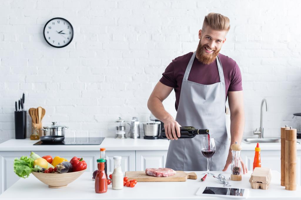 bel giovane barbuto in grembiule versando vino e sorridendo alla macchina fotografica mentre cucinava in cucina
 - Foto, immagini