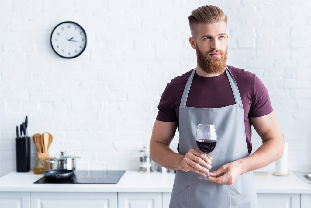 schöner bärtiger junger Mann in Schürze hält ein Glas Rotwein in der Hand und schaut in der Küche weg - Foto, Bild