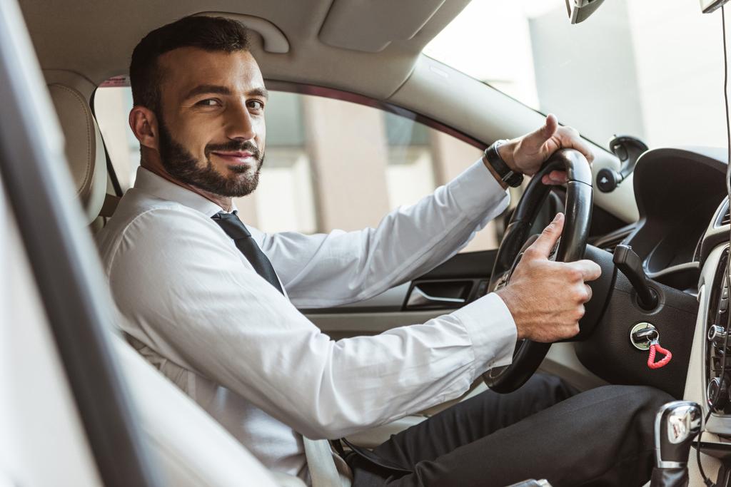 улыбчивый красивый водитель держит руль и смотрит на камеру в машине
 - Фото, изображение