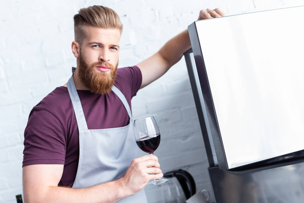 handsome bearded man in apron holding glass of red wine and looking at camera - Photo, Image