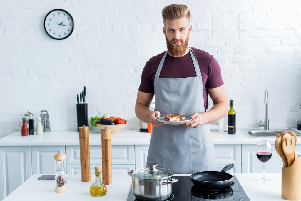 handsome bearded young man in apron holding delicious beef steak on plate and looking at camera - Photo, Image