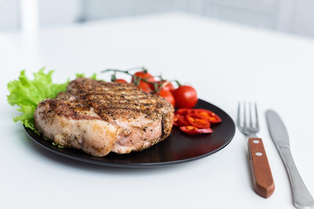 Nahaufnahme von leckeren gegrillten Steaks mit Salat, Pfeffer und Kirschtomaten auf schwarzem Teller - Foto, Bild