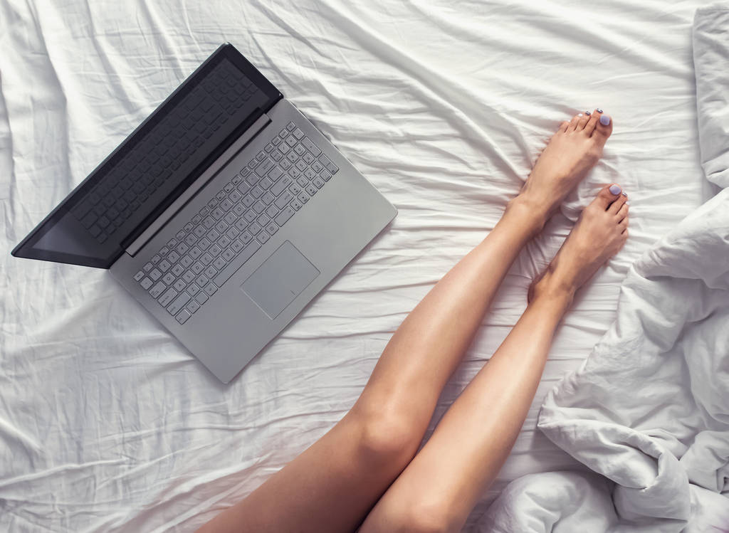 Pernas femininas na cama e laptop de pé perto
. - Foto, Imagem