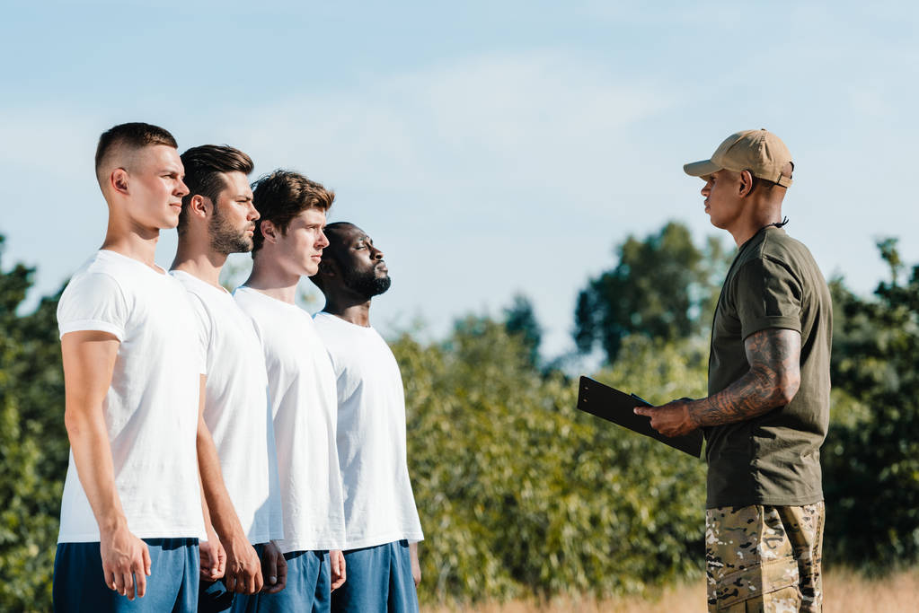 boční pohled na africké americké taktické instruktor s Poznámkový blok a mnohonárodnostní mladých vojáků na rozsah - Fotografie, Obrázek