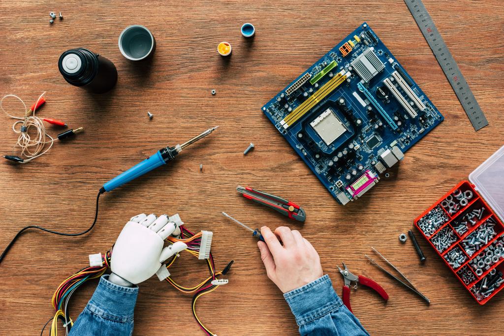 cropped image of man with prosthetic arm holding wires and screwdriver surrounded by instruments on wooden table - Photo, Image