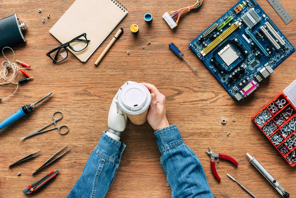 обрезанное изображение инженера-электронщика с протезной рукой, держащей бумажную чашку на столе с инструментами
 - Фото, изображение